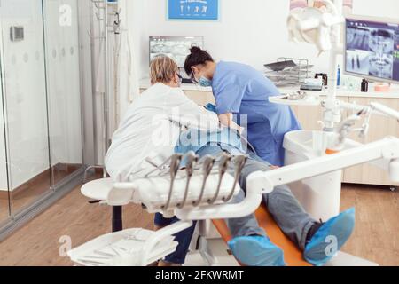 Homme malade assis en chaise dentaire à bouche ouverte pour l'exémination, dans le bureau stomatologique, médecin se préparant pour des problèmes buccaux. Dentiste femme senior et infirmière médicale avec des masques enlève la dent Banque D'Images