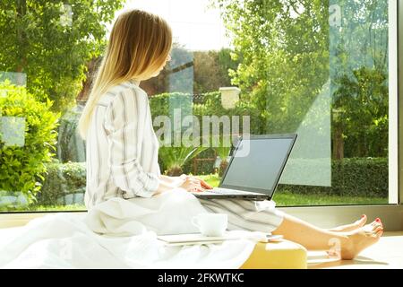 Jeune femme attrayante assise sur le sol, table basse près de la grande fenêtre de vue de jardin, travail, navigation, surf, blogging sur ordinateur portable, vide Banque D'Images