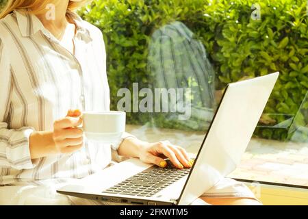 Jeune femme attrayante assise sur le sol près d'une table basse près d'une grande fenêtre avec vue sur le jardin, travail, navigation, surf, blogging sur un ordinateur portable. Perk Banque D'Images