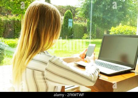 Jeune femme attrayante assise sur le sol, table basse près de la grande fenêtre de vue de jardin, travail, navigation, surf, blogging sur ordinateur portable, vide Banque D'Images