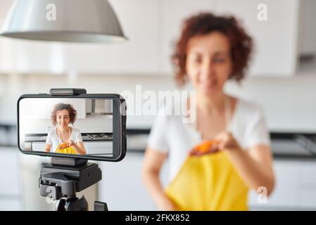 Une jeune femme se enregistre avec son smartphone dans sa cuisine tout en préparant une recette de salade, il y a la lumière naturelle et elle porte un a jaune Banque D'Images