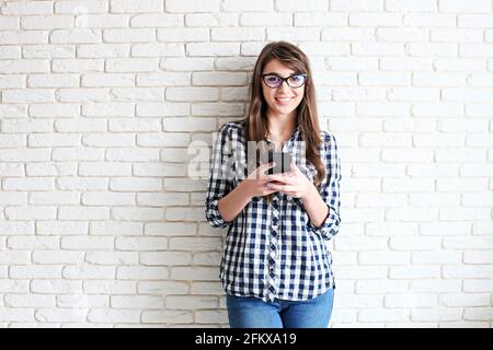 Jeune femme attirante portant une chemise à carreaux et des lunettes pour chat recevant un message sur un nouveau téléphone cellulaire. Jolie femme modèle tenant mobile gadget dans han Banque D'Images