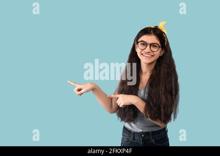 Une jeune fille indienne heureuse pointant à côté de l'espace vide, souriant et regardant l'appareil photo sur fond bleu de studio Banque D'Images