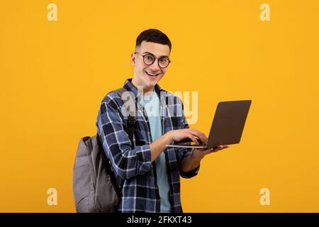 Jeune étudiant intelligent avec sac à dos utilisant un pc portable sur orange arrière-plan du studio Banque D'Images