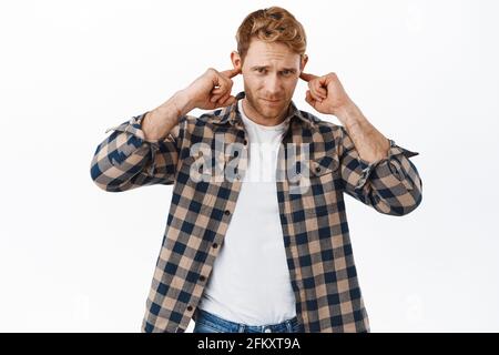 Un homme adulte à tête rouge affligé couvre ses oreilles, branche les doigts dans les trous d'oreille et regarde dérangé par un bruit fort agaçant, ne veut pas entendre quelque chose Banque D'Images