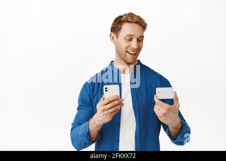 Homme attrayant aux cheveux rouges, payant avec un téléphone mobile et une carte de crédit, en regardant heureux tout en entrant le numéro bancaire dans l'application pour smartphone, faire la livraison Banque D'Images