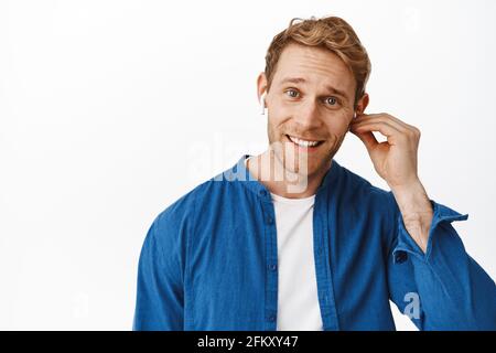 Gros plan de beau redhead homme mettre sur casque sans fil, écouter de la musique ou parler sur téléphone mobile dans des écouteurs, sourire heureux à l'appareil photo, debout Banque D'Images