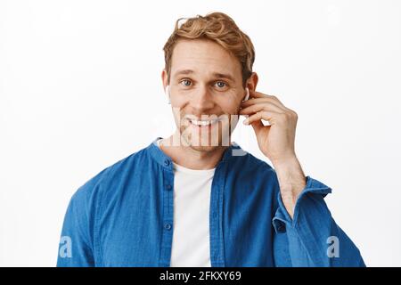 Gros plan sur le beau homme à tête rouge porte des écouteurs sans fil et sourit à l'appareil photo, parle avec quelqu'un dans un casque, écoute de la musique dans des écouteurs, se tient debout Banque D'Images