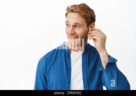 Homme moderne et élégant aux cheveux rouges enfilez un casque sans fil, tournez la tête vers la gauche au logo et souriant heureux, parlant en déplacement, en utilisant des écouteurs pour la vidéo Banque D'Images