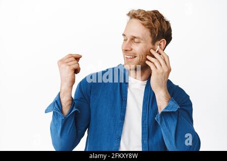 Portrait d'un homme heureux sourit et touche un casque tout en écoutant de la musique, en touchant les doigts et les yeux, en vous détendant avec vos chansons préférées, se dresse sur un blanc Banque D'Images