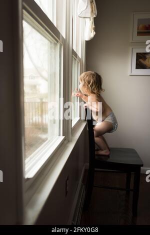 Un jeune enfant dans un couches repose sur une chaise et regarde hors d'une fenêtre Banque D'Images