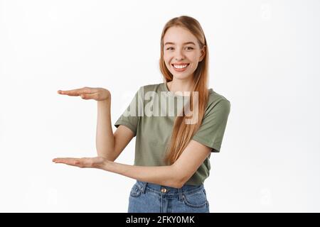 Jeune femme souriante montrant un article, tenant quelque chose, vide entre les mains, regardant heureux, recommandant le produit, debout sur blanc Banque D'Images