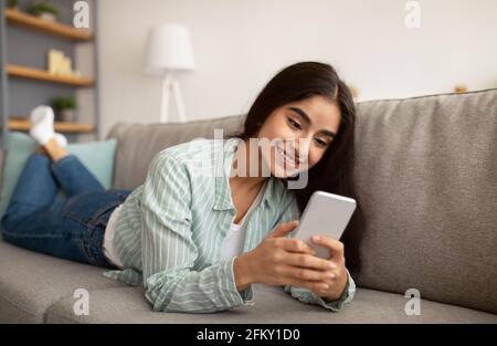 Portrait d'une femme indienne millénaire utilisant un téléphone portable pour travailler ou étudier, allongé sur un canapé à la maison Banque D'Images