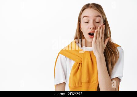 Gros plan d'une jeune fille fatiguée ou ennuyeuse, bâilling avec des yeux fermés, couvrant la bouche ouverte, écoutez quelque chose d'ennuyeux, debout contre un fond blanc Banque D'Images