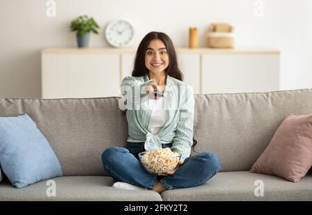 Pleine longueur de jeune femme indienne avec télécommande de regarder Télévision et repas de pop-corn sur le canapé à la maison Banque D'Images
