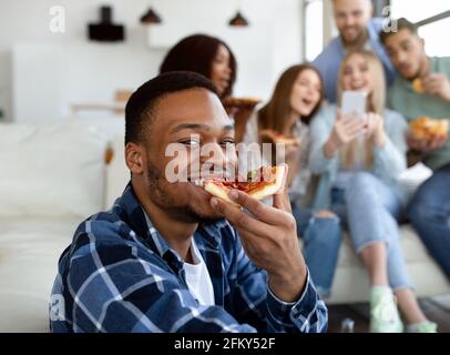 Un gars noir attrayant mangeant une délicieuse pizza, ses divers amis avec un téléphone portable ayant du plaisir sur le fond Banque D'Images