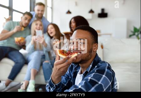 Beau gars noir mangeant une tranche de pizza, groupe de ses amis multiethniques avec smartphone sur fond Banque D'Images
