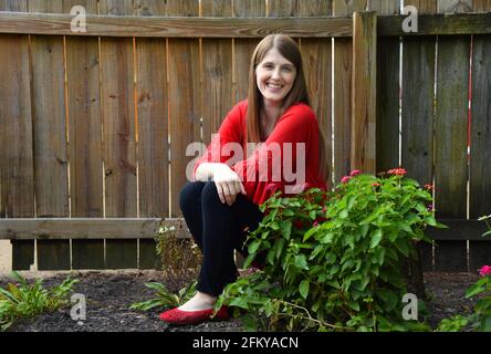 Une jeune femme se détend dans son jardin. La clôture rustique en bois est en arrière-plan et les fleurs sont en fleurs. Elle porte un pantalon noir et un chemisier rouge. Banque D'Images