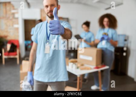 Homme volontaire en uniforme bleu et gants de protection montrant les pouces vers le haut à l'appareil photo. Tri en équipe, emballage des articles dans des boîtes en carton Banque D'Images