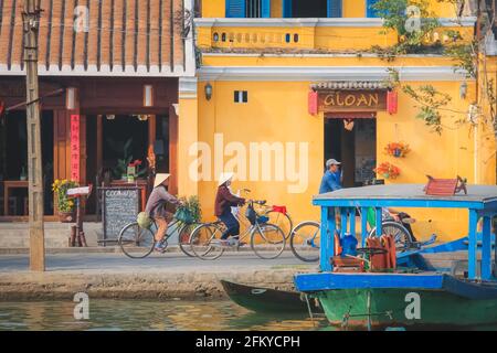 Hoi an, Vietnam - Mars 6 2019: Les cyclistes vietnamiens locaux le long de la rivière Thu bon et vibrant, jaune bâtiments traditionnels et rues de H Banque D'Images