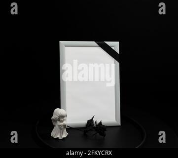 Cadre blanc avec ruban de deuil noir, rose noire et fond d'ange. Image funéraire conceptuelle Banque D'Images