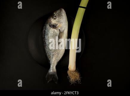 Un poisson cru de la dorade dans une assiette noire sur Arrière-plan noir avec leek Dark Mood photographie Banque D'Images