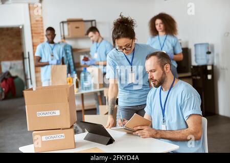 Équipe de bénévoles divers en uniforme bleu prenant des notes, calculant, triant et emballant des articles dans des boîtes en carton tout en travaillant ensemble sur le don Banque D'Images