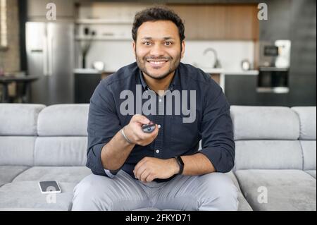 Un jeune indien enthousiaste regardant des émissions de télévision, assis sur le canapé confortable avec télécommande, changer de chaîne, regarde la caméra, un gars multiracial passe des loisirs à la maison Banque D'Images