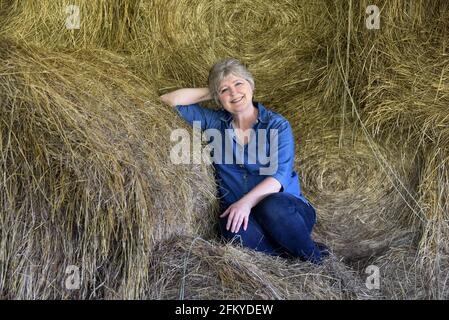 La femme mûre se détend dans le foin. Elle porte un Jean et une chemise en denim. Son sourire dit tout, la coupe de foin est terminée. Banque D'Images