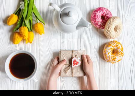 Les mains de l'enfant tiennent le cadeau dans un emballage fait. Beignets, tulipes et bouilloire sur table. Romantique Saint Valentin petit déjeuner au lit pour votre cher. c Banque D'Images