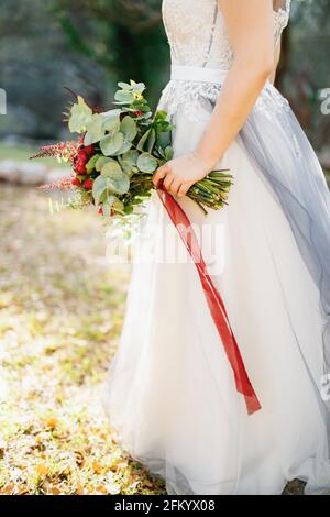 Une mariée dans une robe de mariage gris pâle tient un bouquet de pivoines rouges, astilba, roses et eringium et de longs rubans rouges, gros plan Banque D'Images