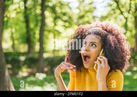 Jolie femme afro parlant sur le téléphone portable dans l'étonnement, dans un jardin. Banque D'Images