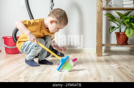 Un petit garçon mignon aide à nettoyer la maison par balayer les ordures avec un balai d'enfant du sol dans une pelle à poussière en peluche Banque D'Images