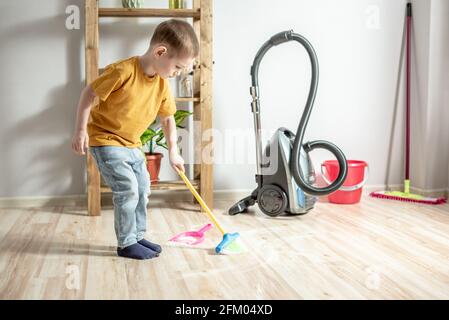 Un petit garçon mignon aide à nettoyer la maison par balayer les ordures avec un balai d'enfant du sol dans une pelle à poussière en peluche Banque D'Images