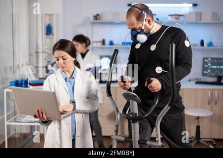 Homme athlète courant sur un entraîneur de croix avec des électrodes attachées à son corps et son masque. Le médecin utiliserun ordinateur portable et contrôle les données EKG affichées sur les moniteurs de laboratoire, discutant avec le patient. Banque D'Images