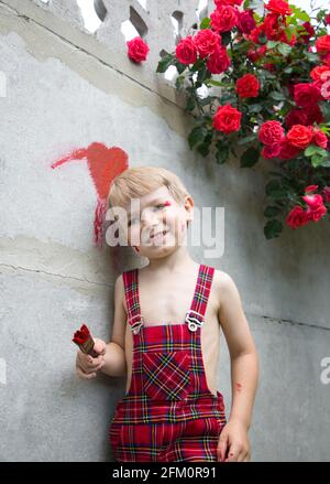 mignon garçon dans une combinaison à carreaux peint un coeur sur la clôture avec de la peinture rouge. Derrière lui se trouve une fleur de roses rouges. Saint-Valentin, un cadeau à faire Banque D'Images