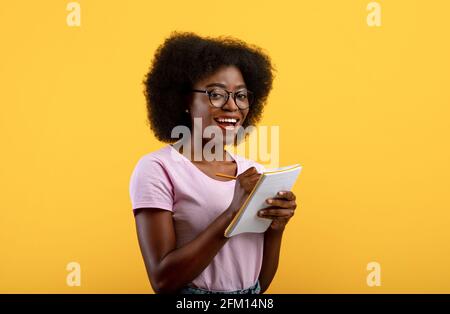 Concept de liste de contrôle. Smart noire étudiante vêtue de lunettes, prenant des notes dans le livre sur fond jaune studio Banque D'Images