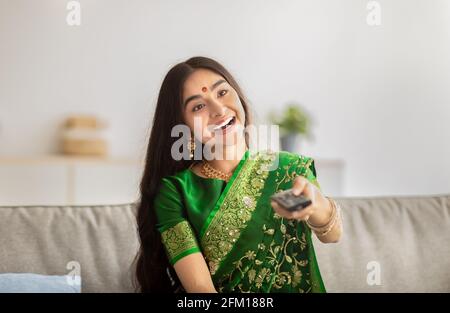 Portrait d'une jeune femme indienne en sari traditionnel tenant la télécommande, regardant la télévision à la maison Banque D'Images