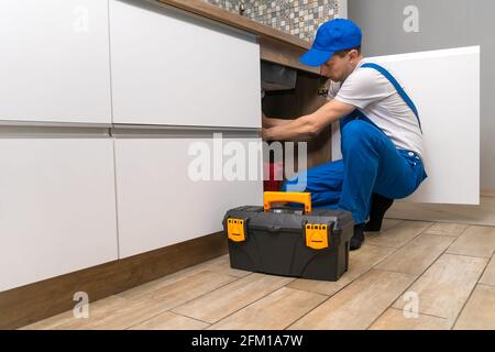 Le handyman en combinaison répare les tuyaux sous l'évier dans la cuisine, à côté d'une boîte d'outils Banque D'Images