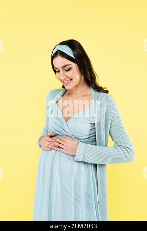 femme enceinte joyeuse tenant les mains sur le ventre tout en souriant isolé sur jaune Banque D'Images