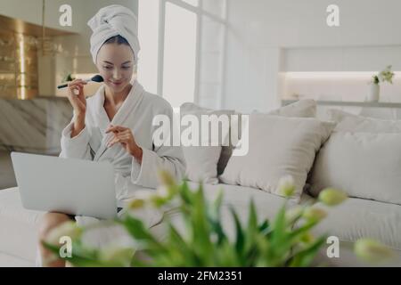 Jeune jolie femme en peignoir faisant le maquillage quotidien tout en regardant didacticiel vidéo sur ordinateur portable Banque D'Images