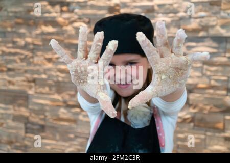 Une jeune fille souriante, vêtue comme le montre un chef professionnel sa farine et sa pâte se tachèrent les mains Banque D'Images