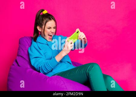 Photo de jeune fou criant gai fille assis fauteuil jouer jeux vidéo dans le téléphone isolés sur fond rose Banque D'Images