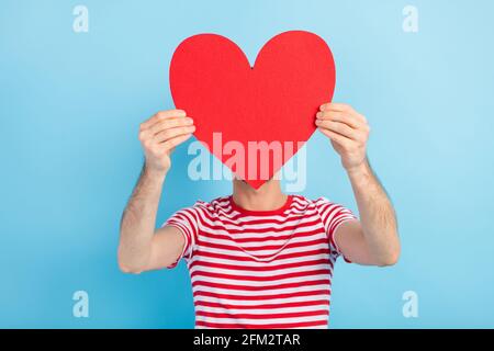 Photo de doux timide jeune homme habillé t-shirt rayé rouge papier coeur couverture face tête vide espace isolé couleur bleue arrière-plan Banque D'Images