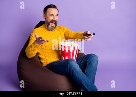 Photo d'un homme assis, sac à bandoulière, boîte de commande à distance, pop-corn portez un jean chandail jaune sur fond violet isolé Banque D'Images