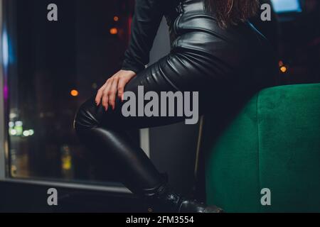 Belle jeune femme assise sur un fauteuil en cuir avec repose-pied. Femme portant un pantalon et des bottes en cuir noir à l'intérieur. Photo noir et blanc avec couleur Banque D'Images