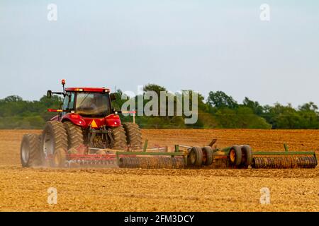 Un tracteur à six roues rouge avec cabine laboure un champ agricole vierge au printemps. L'outil de charrue fixé crée un nuage de poussière sur la ferme. IMAG Banque D'Images