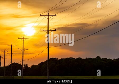 Silhouettes de poteaux électriques transportant de l'électricité haute tension le long d'une route dans une zone rurale. Coucher de soleil, lever de soleil avec ciel nuageux orange rouge et Banque D'Images