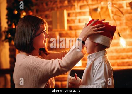 La jeune mère met le chapeau de Noël du Père Noël sur son enfant. Tasse de cacao avec guimauves dans la main du garçon. Boîtes-cadeaux, guirlandes et branches de sapin sur un mur en brique. Nouvel an. Vacances d'hiver. Banque D'Images