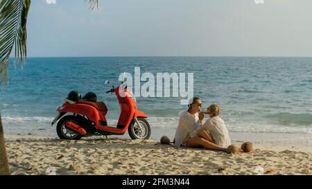 Trajet en scooter. Un couple charmant sur une moto rouge vêque de vêtements blancs sur une plage de sable. Les gens qui marchent près des palmiers tropicaux, de la mer. Location de motos. Banque D'Images